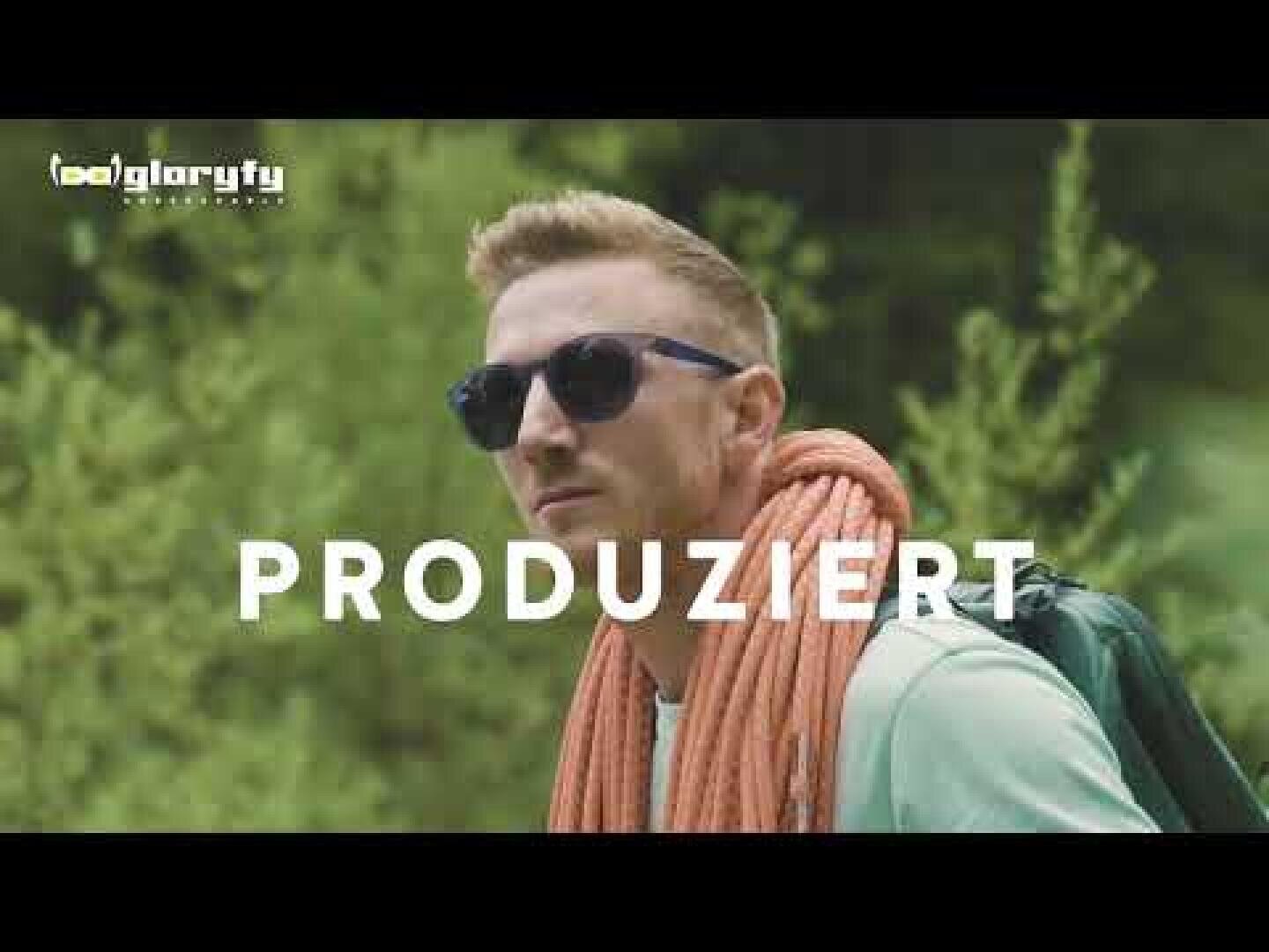 Ein Mann mit Sonnenbrille und hellgrünem Hemd steht im Freien mit einem aufgerollten orangefarbenen Seil über der Schulter und einem Rucksack. Der Hintergrund ist grün und belaubt. Das Wort PRODUZIERT ist in weißer Schrift überlagert.