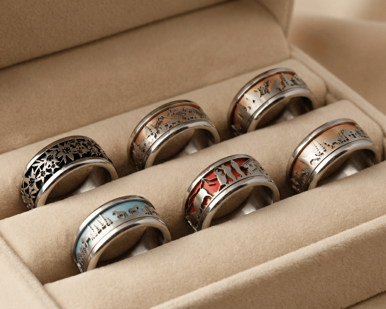 Sechs dekorative Ringe in einer beigen Samtschachtel, jeder mit einzigartigen Gravurmustern und verschiedenfarbigen Einlagen in Schwarz, Blau, Rot und Brauntönen.