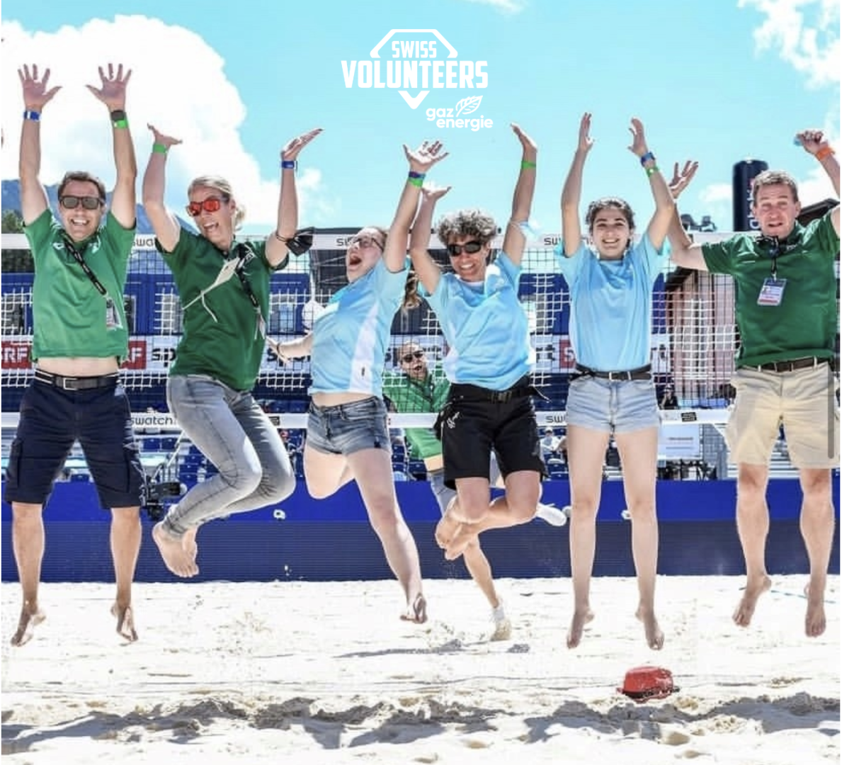 Sechs Personen an einem Sandstrand springen in die Luft, lächeln und tragen Freiwilligen-Shirts. Hinter ihnen sind ein Netz und ein blauer Himmel zu sehen. Das Logo von Swiss Volunteers ist oben auf dem Bild zu sehen.