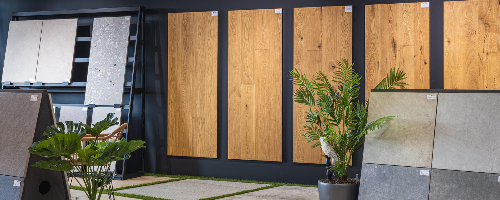 In einem Ausstellungsraum werden große Muster von Holz- und Steinfliesen an den Wänden ausgestellt, mit Grünpflanzen im Vordergrund und weiteren Mustern von Bodenfliesen darunter.