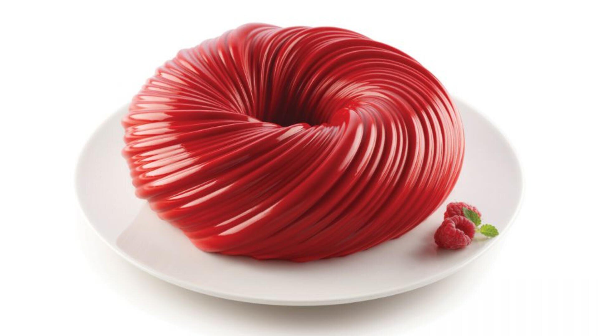 Auf einem weißen Teller liegt ein glänzender, roter Bundt-Kuchen mit Marmormuster, daneben liegen zwei Himbeeren und ein Minzezweig.