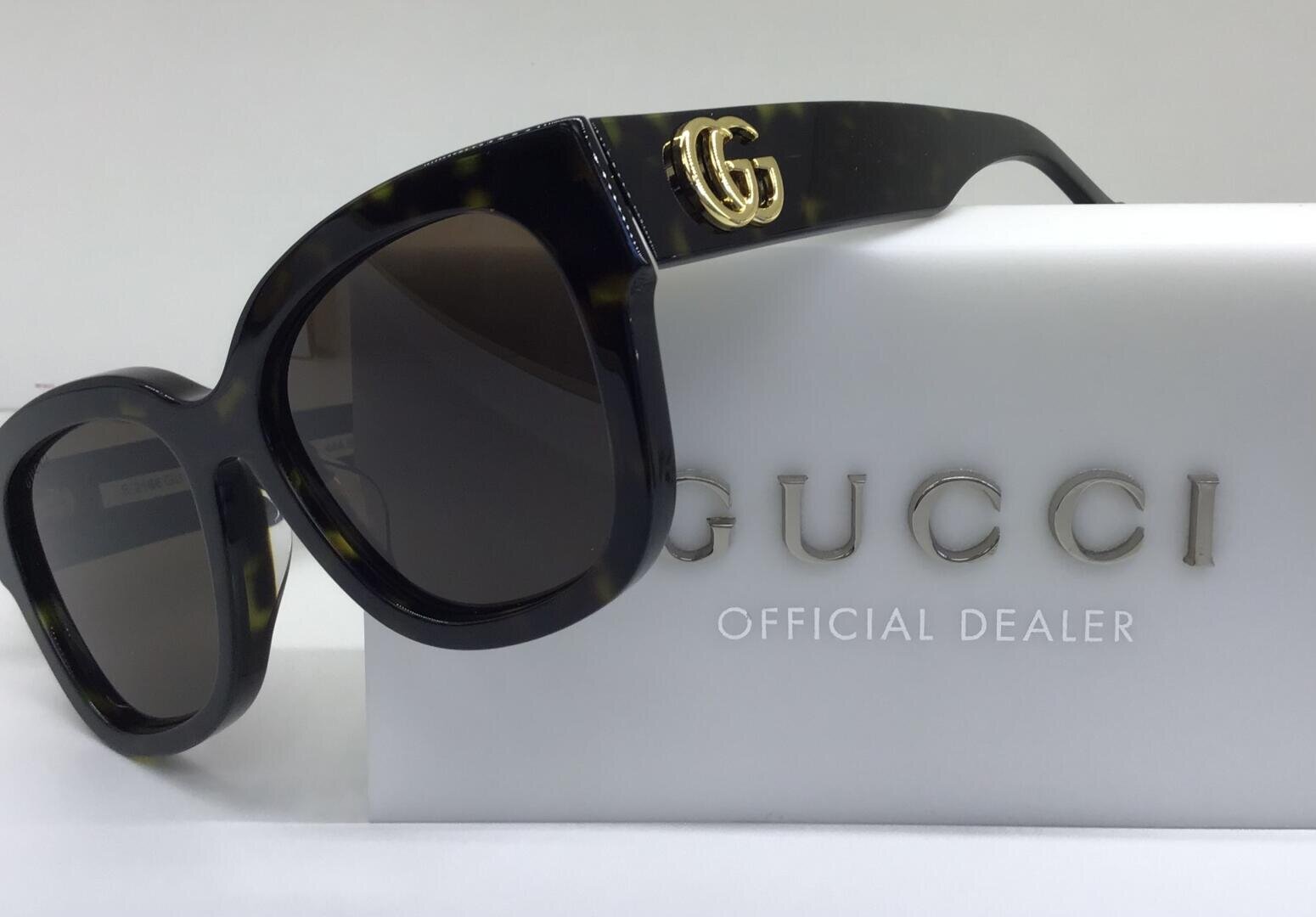 Eine schwarze Gucci-Sonnenbrille mit goldenem GG-Logo an der Seite ruht auf einer weißen Box mit dem Gucci-Markennamen und dem Schriftzug Official Dealer in silberner Schrift.