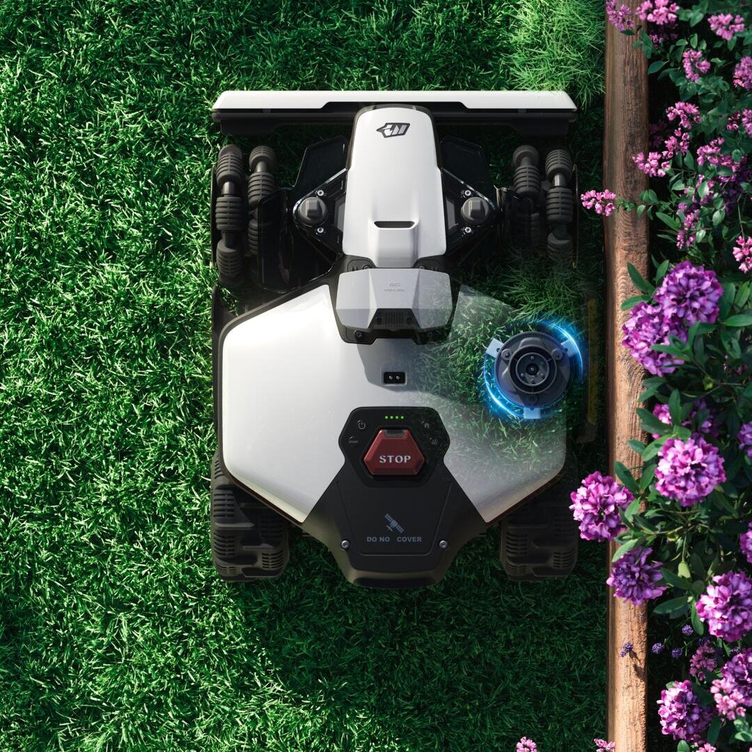Ein Rasenmähroboter mäht grünes Gras in der Nähe eines hölzernen Blumenbeetes mit blühenden lila Blumen. Der Mäher hat ein schlankes, modernes Design und eine auffällige rote Stopptaste auf seiner Oberfläche.