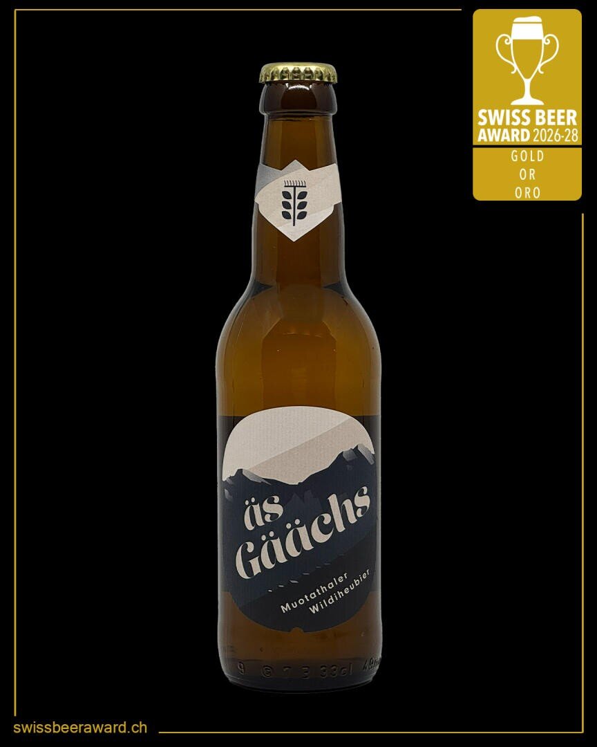 Eine Bierflasche aus braunem Glas mit der Aufschrift "üs Gäälchs" und einem Bergmotiv auf dem Etikett. In der rechten oberen Ecke befindet sich eine goldene Plakette mit der Aufschrift "SWISS BEER AWARD 2026-28 GOLD OR ORO". Website: swissbeeraward.ch.