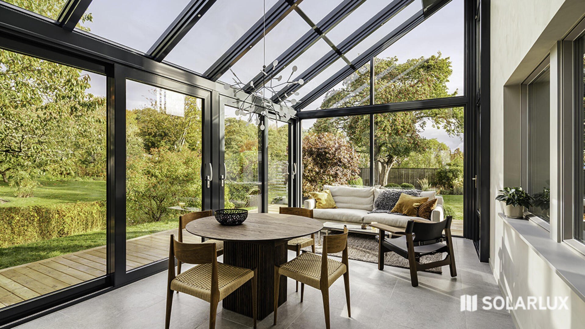 Ein modernes Sonnenzimmer mit großen Glasfenstern und einer Decke mit einem runden Holztisch mit vier Stühlen und einem weißen Sofa mit Blick auf einen grünen, baumbestandenen Garten. Das SOLARLUX-Logo befindet sich in der unteren rechten Ecke.