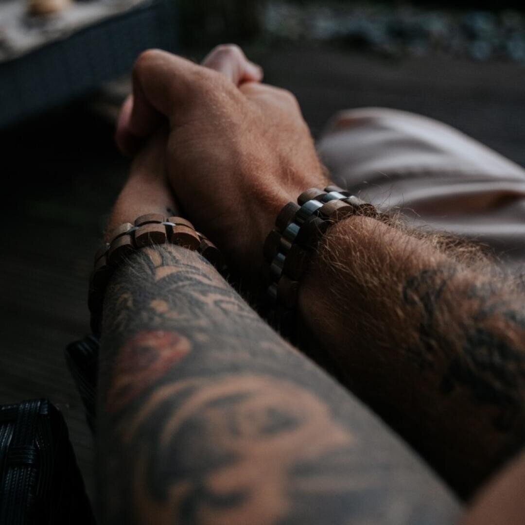 Zwei tätowierte Arme mit Armbändern sind in einem sanften Händedruck verschränkt, der Trost oder Zuneigung suggeriert. Das Bild ist eng auf die Hände und Arme fokussiert, der Hintergrund ist unscharf.