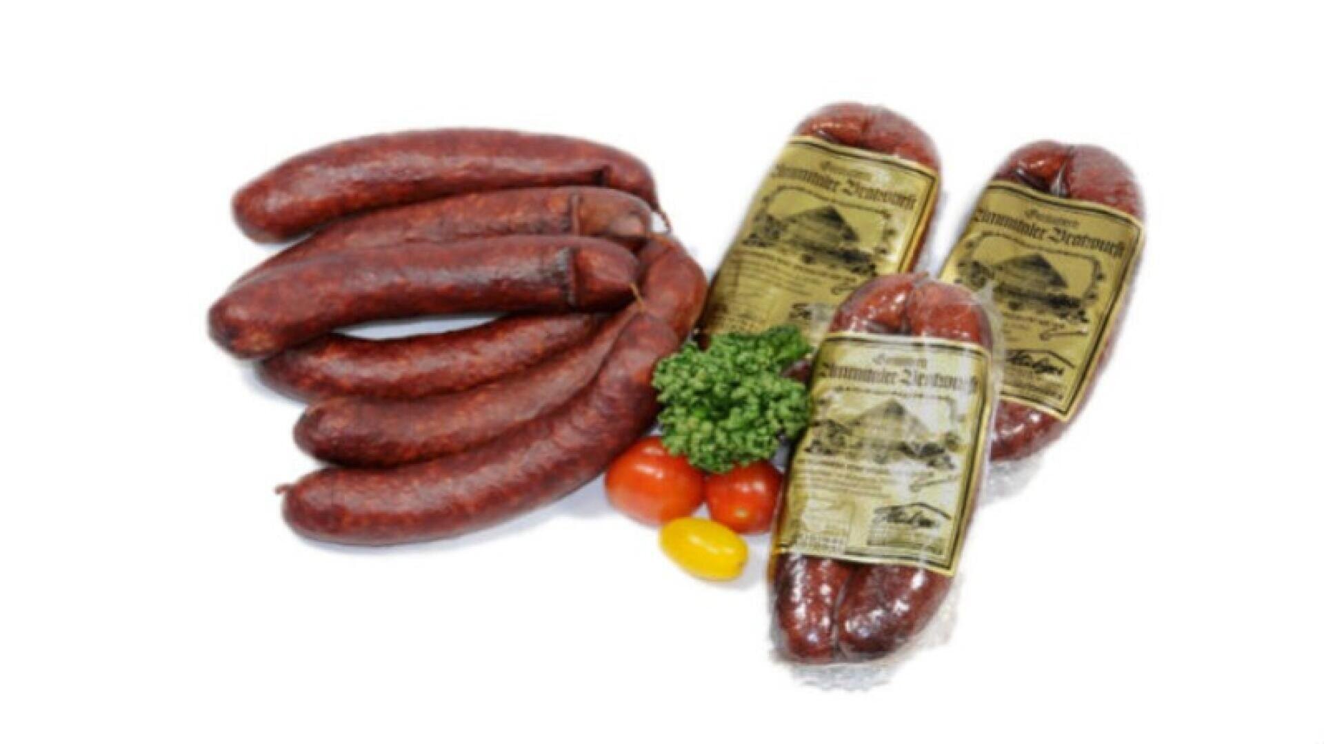 Mehrere geräucherte Würste und drei vakuumverpackte Würste mit Etiketten sind auf einer weißen Fläche angeordnet, begleitet von Petersilie, zwei roten Tomaten und einer gelben Tomate.