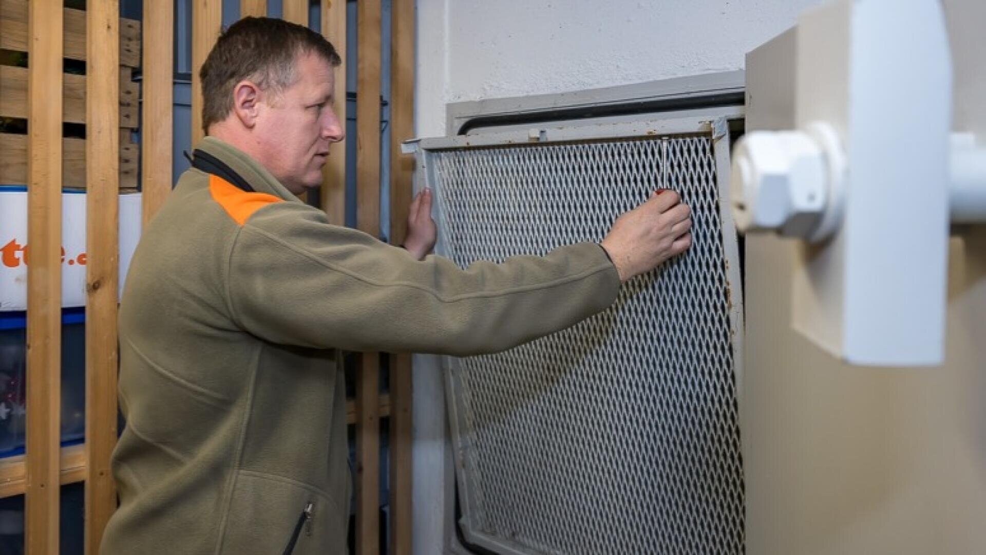 Ein Mann in grüner Jacke ist dabei, ein Metallgitter über einer großen Entlüftung oder Öffnung in einem Industrie- oder Wirtschaftsraum anzubringen oder zu befestigen. In der Nähe sind Holzlatten und eine weiße Maschine zu sehen.