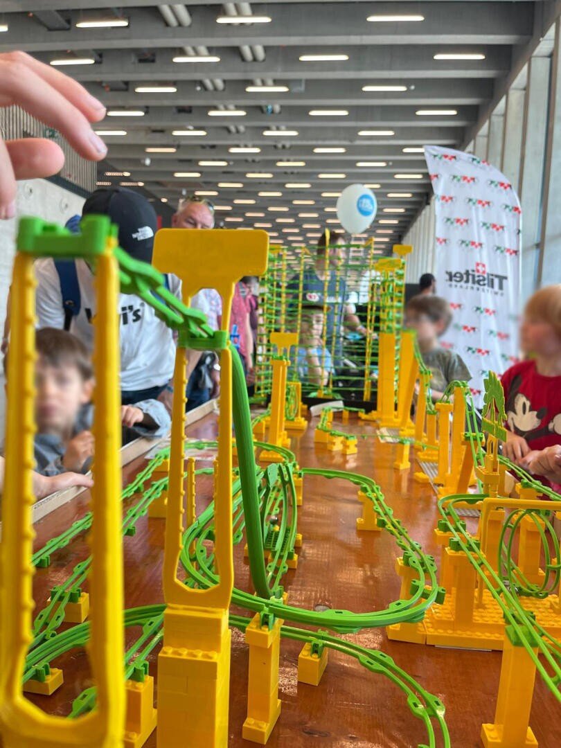 Kinder und Erwachsene versammeln sich um einen Tisch mit einer großen, verschlungenen gelben und grünen Spielzeug-Achterbahnschiene, bauen und spielen. Der Hintergrund zeigt einen modernen Innenraum mit Balken und Fenstern. Die Gesichter sind zum Schutz der Privatsphäre unscharf.