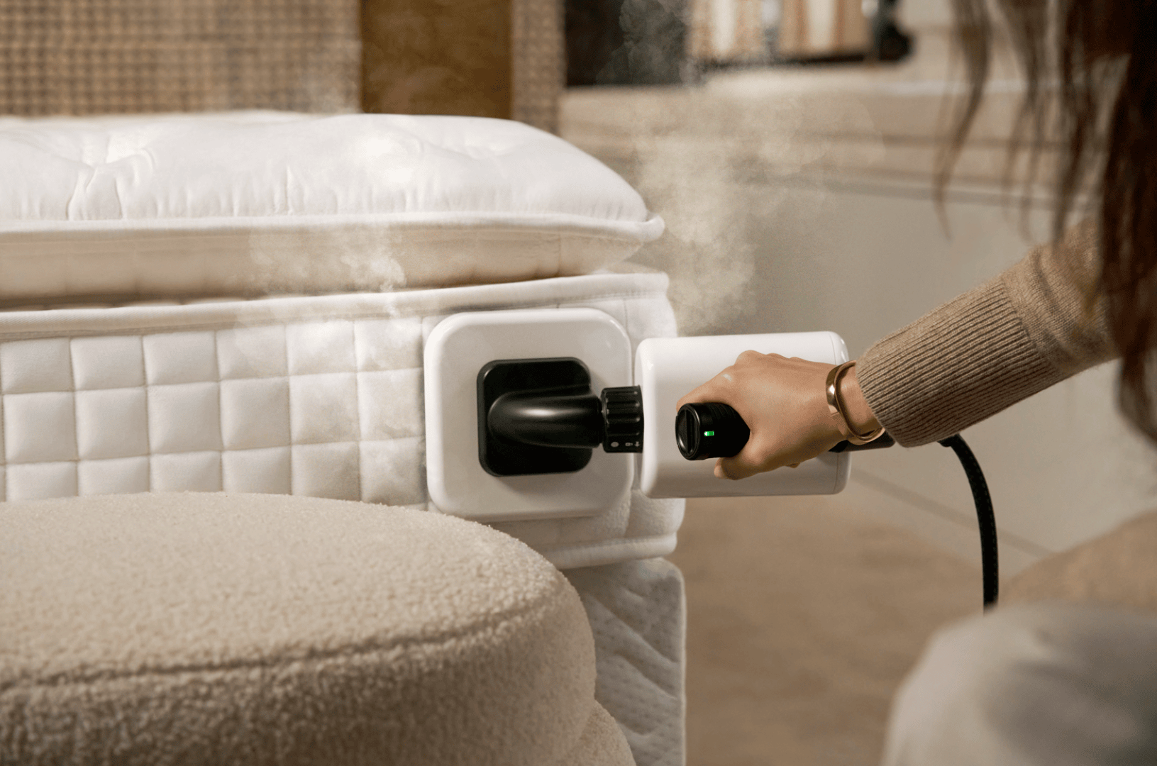 Eine Person hält ein Gerät in der Hand, das an einen Anschluss an der Seite einer weißen Matratze angeschlossen ist, aus der sichtbarer Dampf aufsteigt, was darauf hindeutet, dass die Matratze desinfiziert oder gereinigt wird.