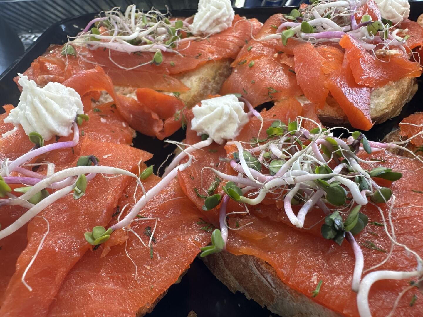 Getoastete Brotscheiben, belegt mit leuchtend orangefarbenem Räucherlachs, Frischkäse und frischen grünen Sprossen, angerichtet auf einem dunklen Tablett.