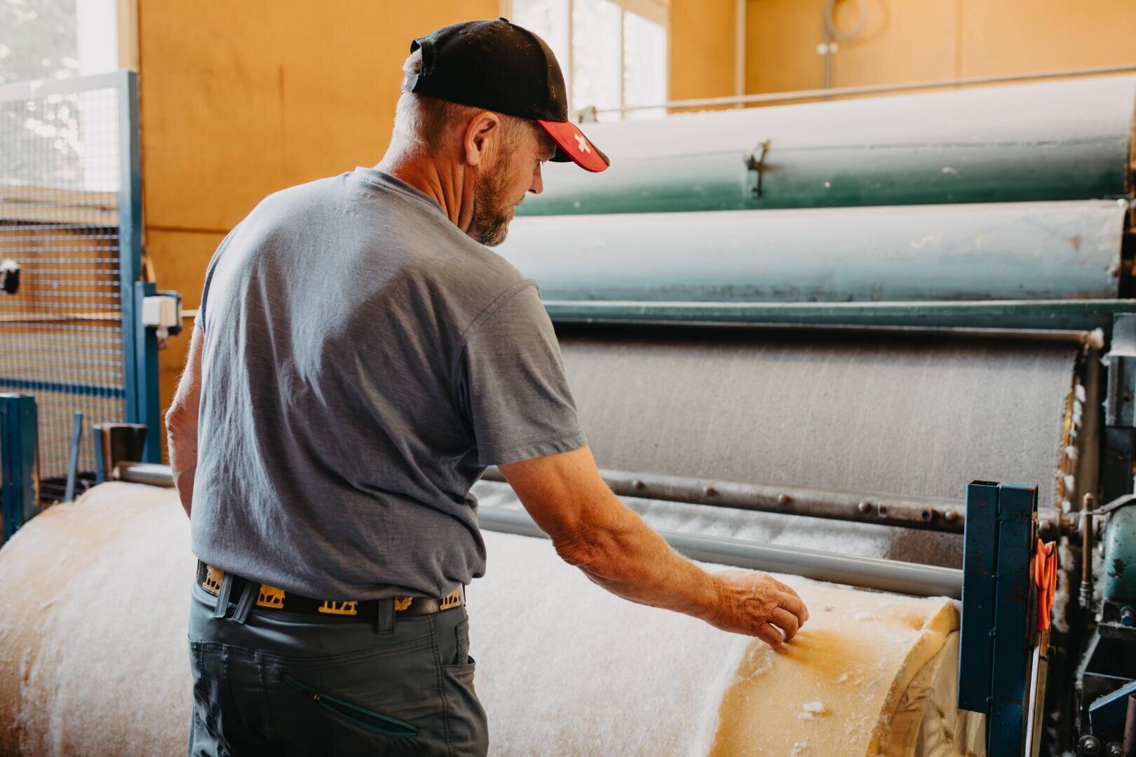 Ein Mann mit Mütze und grauem Hemd inspiziert eine große Rolle gelben Stoffs oder Materials in einer Fabrikhalle, die neben Industriemaschinen steht.