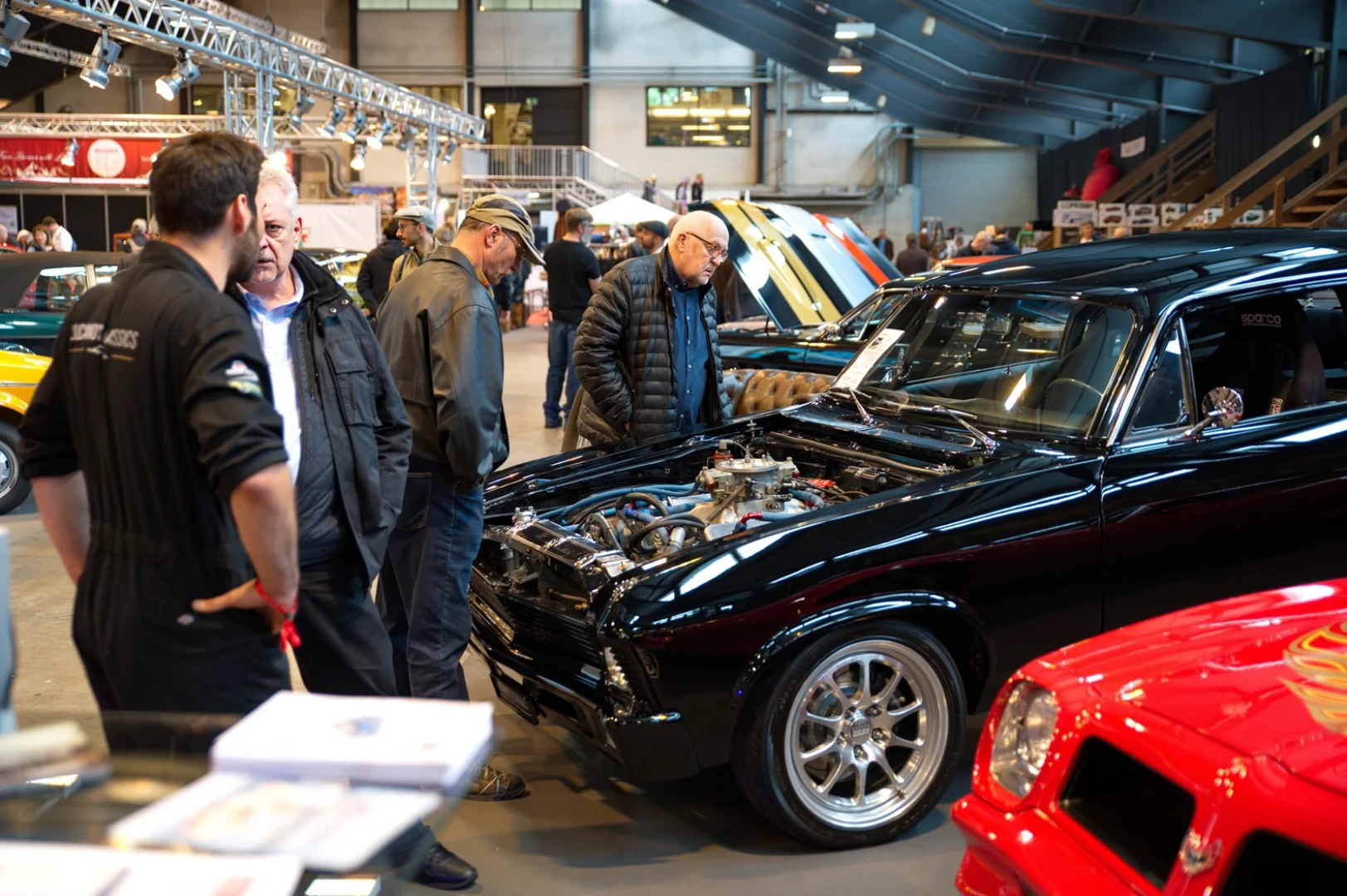 Menschen bewundern Oldtimer auf einer Indoor-Autoshow. Mehrere Männer stehen um ein glänzend schwarzes Auto mit geöffneter Motorhaube und präsentieren den Motor. Im Hintergrund sind weitere farbenfrohe Oldtimer und Ausstellungsstücke zu sehen.