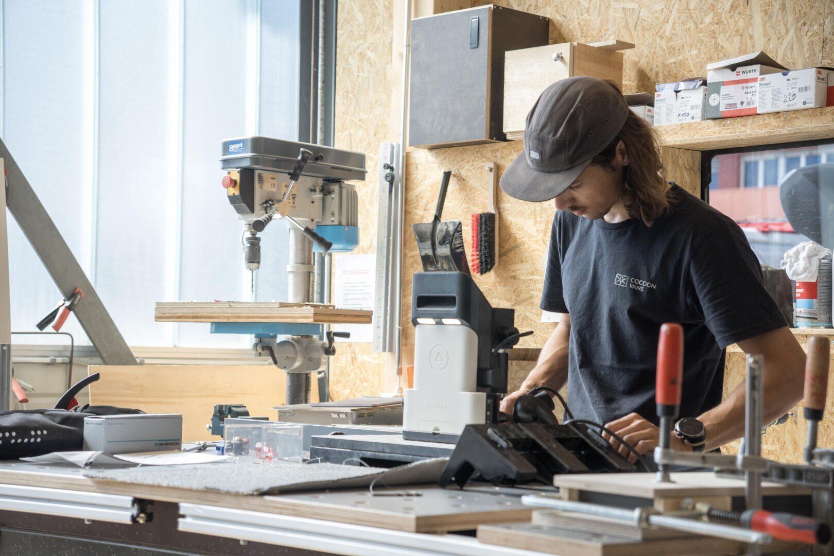 Eine Person mit einer Mütze bedient Maschinen in einer Werkstatt, umgeben von Werkzeugen, Klemmen und Geräten, wobei das Sonnenlicht durch ein Fenster im Hintergrund fällt.