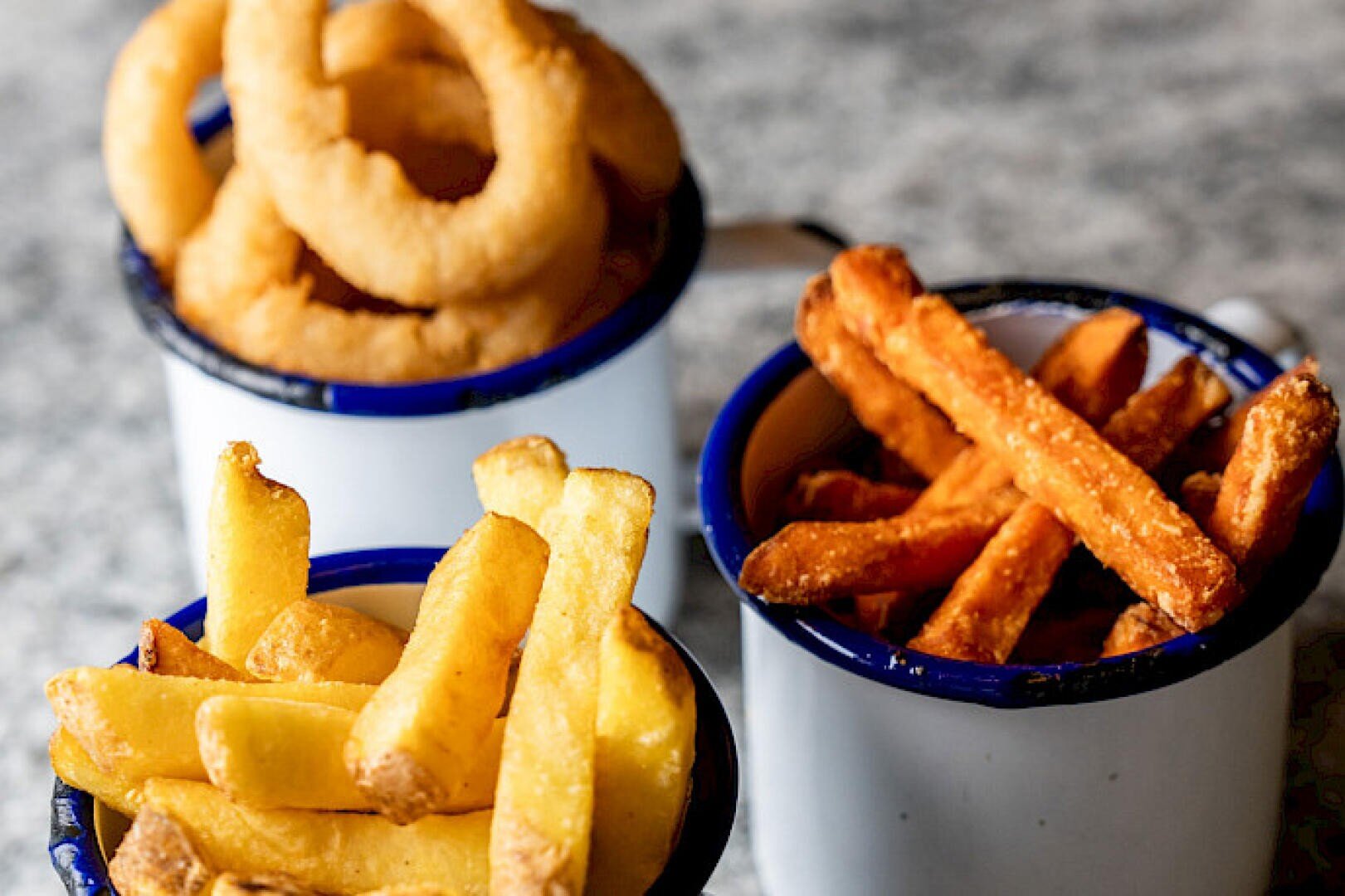 Drei mit Snacks gefüllte Emailleschalen: eine mit goldenen Zwiebelringen, eine mit knusprigen Süßkartoffelpommes und eine mit klassischen Pommes frites, die alle auf einer grauen Fläche stehen.