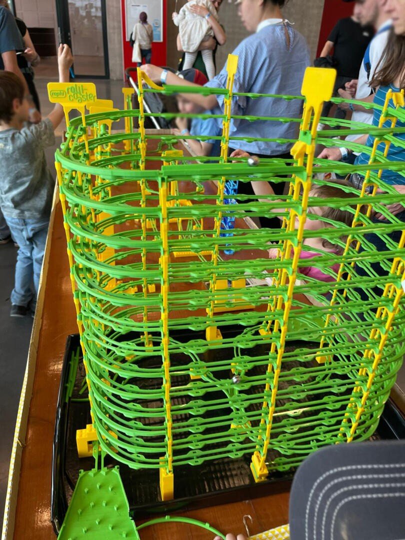Eine Gruppe von Kindern und Erwachsenen interagiert bei einer Indoor-Veranstaltung mit einer großen, grün-gelben Murmelbahn. Die Murmelbahn hat gewundene Bahnen und ist auf einem Holztisch aufgebaut.