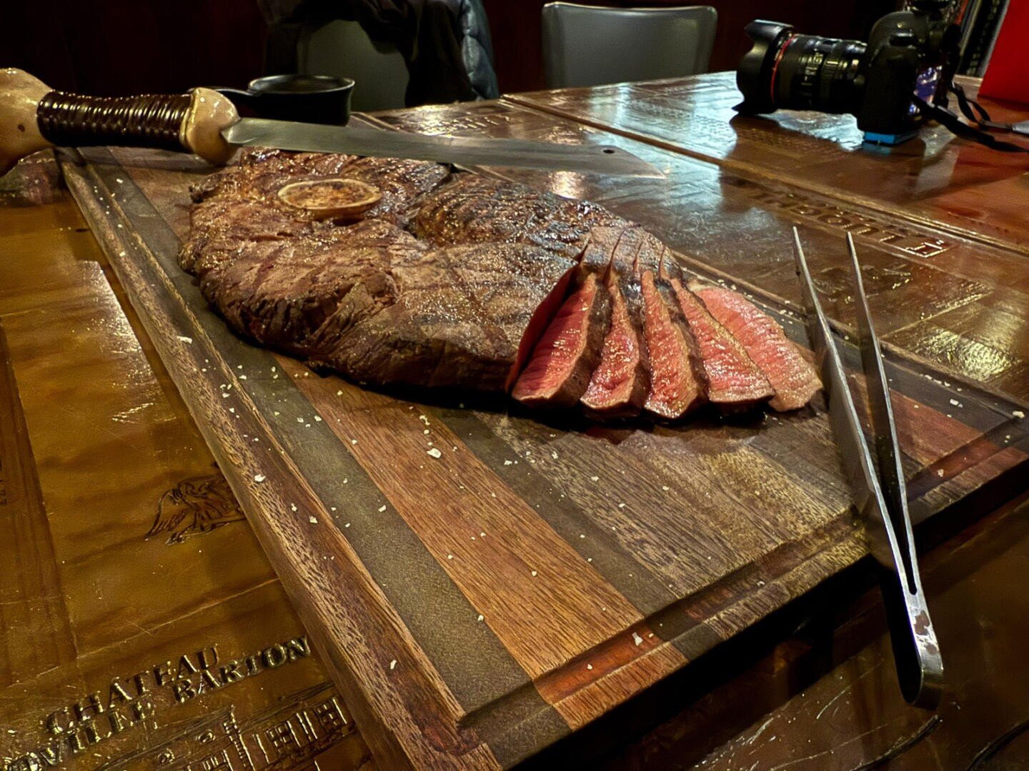 Ein großes, in Scheiben geschnittenes Steak liegt auf einem hölzernen Schneidebrett, das Tranchierwerkzeug liegt daneben. Das Steak ist medium rare und saftig, mit einer gebräunten Kruste. Auf dem Holztisch im Hintergrund befindet sich eine Kamera.