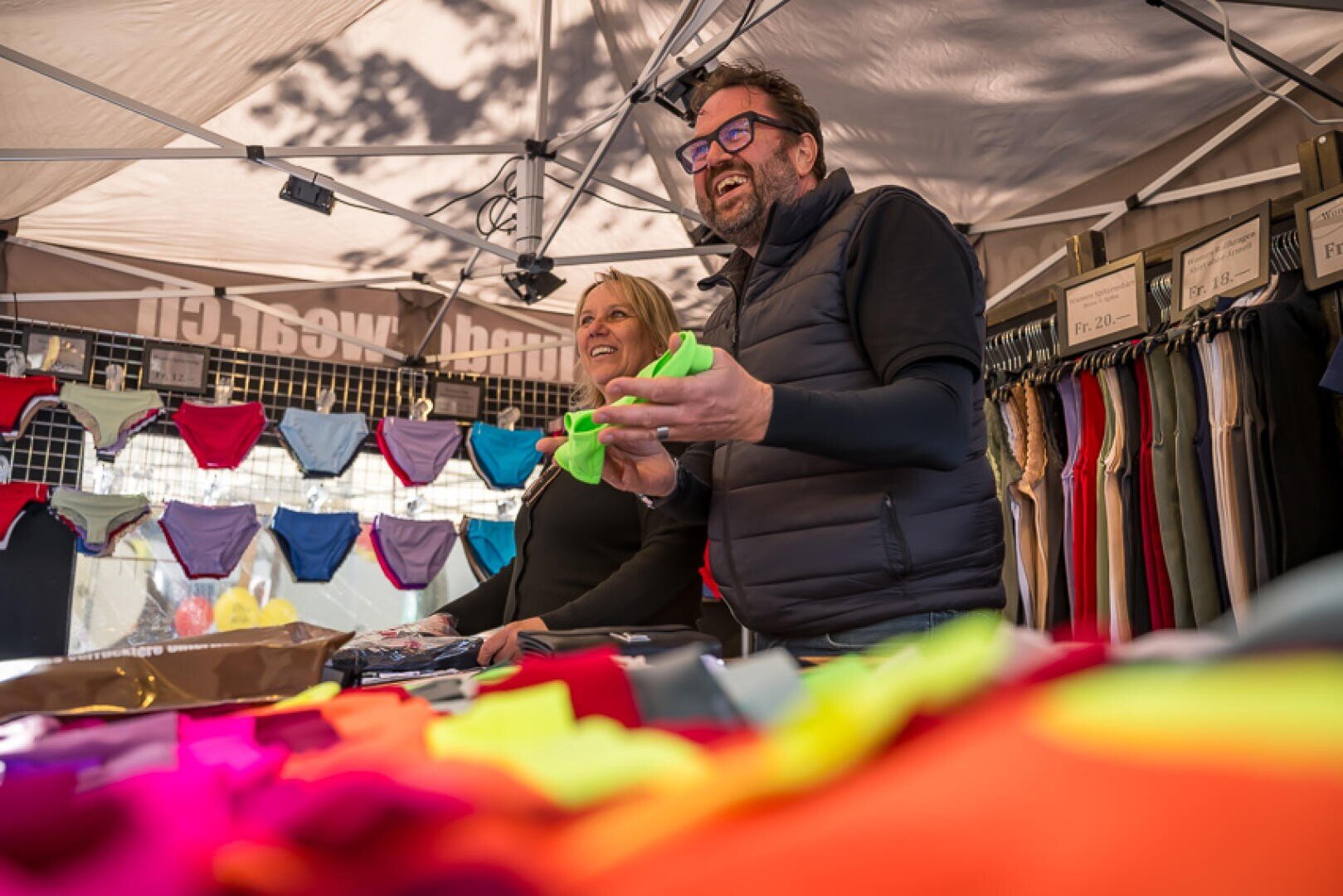 Zwei lächelnde Verkäufer stehen unter einem Vordach eines Marktstandes und präsentieren und verkaufen bunte Unterwäsche. Der Vordergrund ist mit heller Kleidung gefüllt, und hinter ihnen hängen Regale mit Kleidungsstücken. Beide wirken engagiert und glücklich.