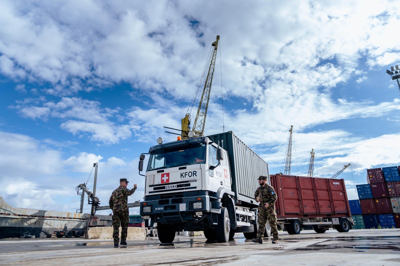 Drei uniformierte Soldaten stehen in der Nähe eines weißen KFOR-Militärlastwagens mit einem medizinischen Symbol in einem Verschiffungshafen, umgeben von Schiffscontainern und Kränen unter einem teilweise bewölkten Himmel.