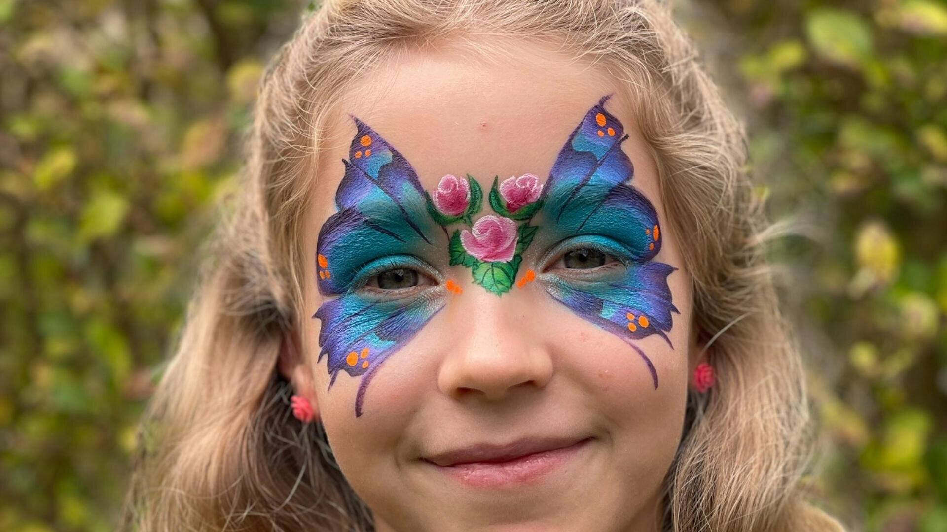 Ein kleines Kind mit langen blonden Haaren lächelt, sein Gesicht ist mit einem bunten Schmetterlingsmuster in Blau und Lila bemalt, das mit rosa Rosen und orangefarbenen Punkten akzentuiert ist.