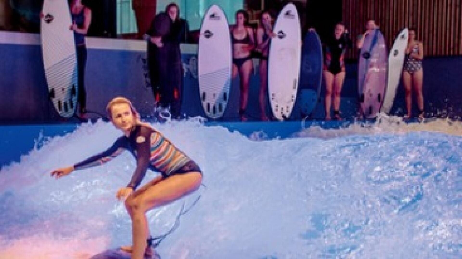 Eine Frau in einem gestreiften Badeanzug surft auf einer Indoor-Welle, während mehrere Personen mit Surfbrettern im Hintergrund zusehen.