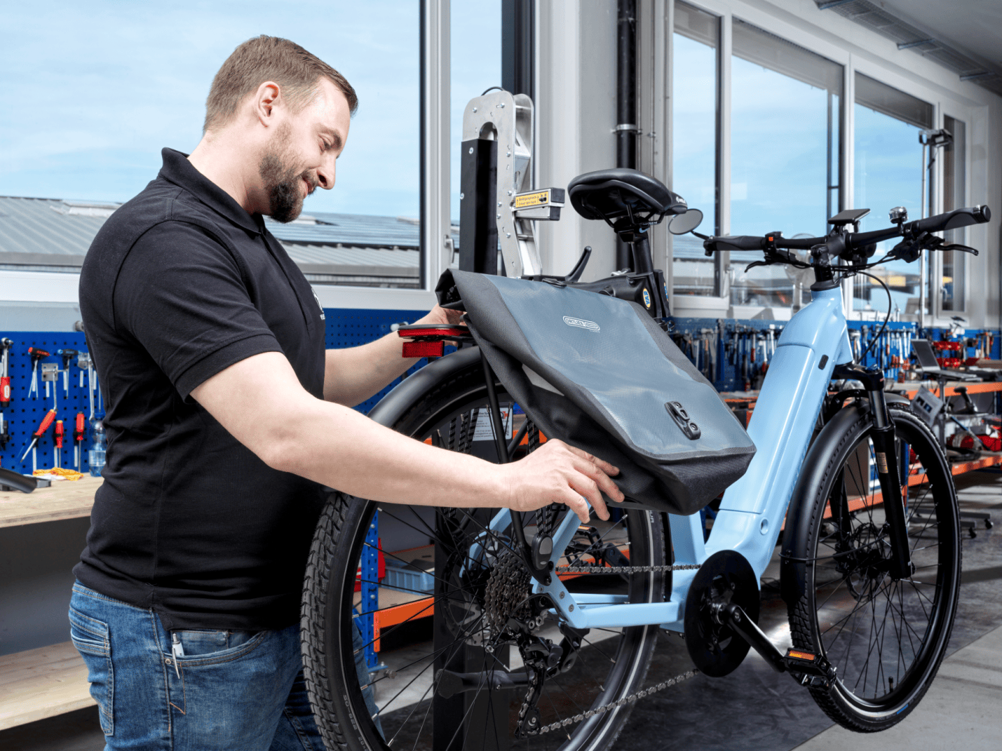 Ein Mann in einem schwarzen Hemd befestigt eine große schwarze Gepäckträgertasche auf dem Gepäckträger eines blauen Elektrofahrrads in einer hellen, modernen Werkstatt mit Werkzeugen und Werkbänken im Hintergrund.