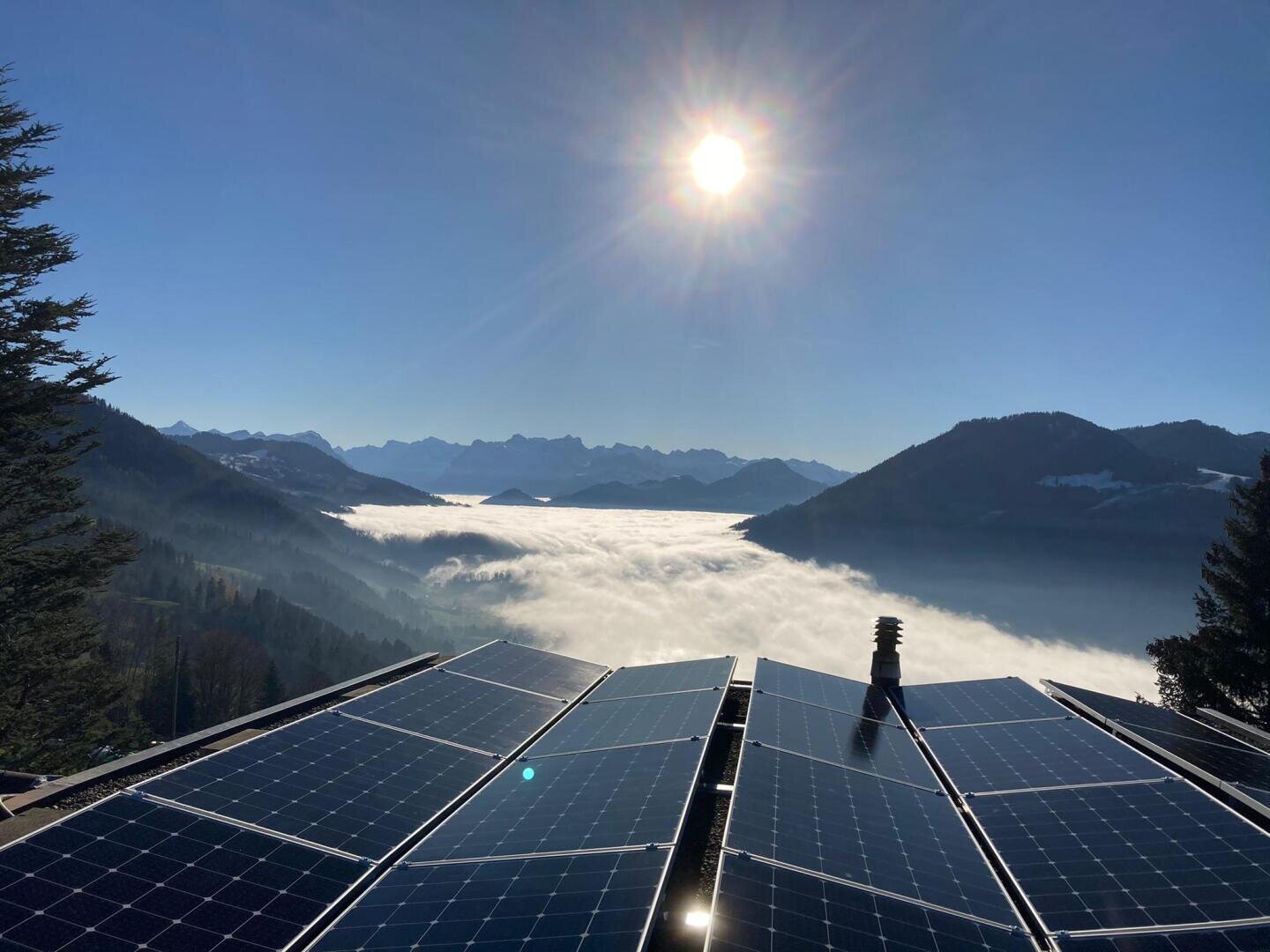 Solarmodule auf einem Dach überblicken ein nebelverhangenes Tal, das von Bergen umgeben ist, und die Sonne strahlt von einem klaren blauen Himmel. Kiefern rahmen die Szene ein, und am Horizont sind ferne Gipfel zu sehen.