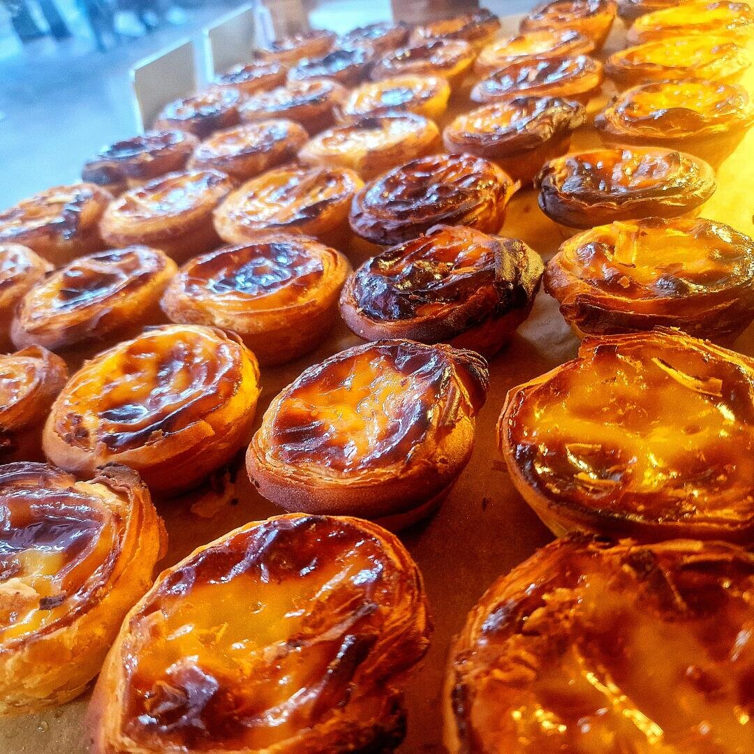 Reihen von goldbraunen portugiesischen Puddingtörtchen (pastéis de nata) mit karamellisierten Oberseiten, die dicht nebeneinander auf einem Tablett angeordnet sind.