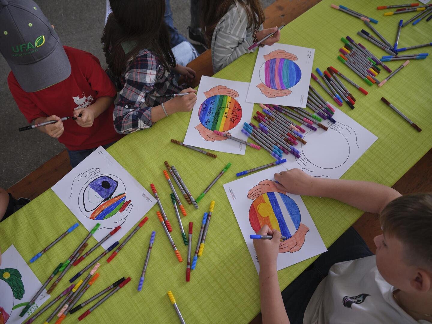 Kinder sitzen um einen Tisch herum und malen mit Stiften Zeichnungen von Händen, die eine Weltkugel halten. Der Tisch ist mit Buntstiften und fertigen bunten Kunstwerken bedeckt.