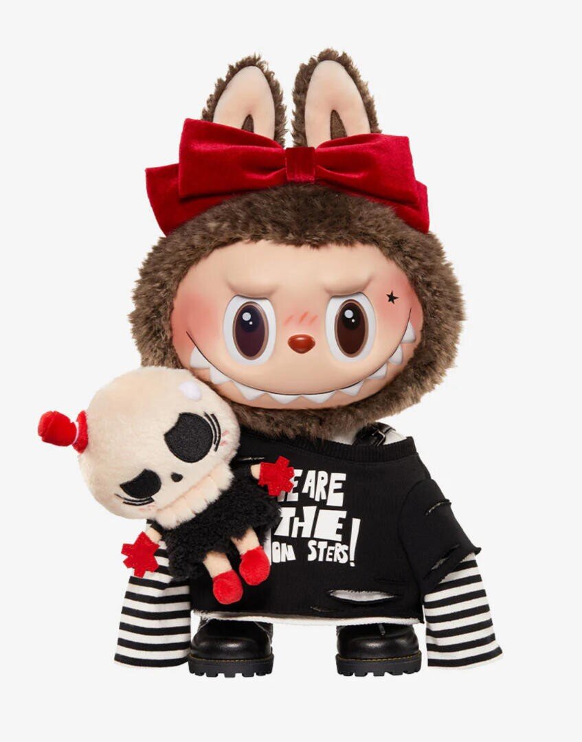 Eine Puppe mit großen Augen, einem verschmitzten Grinsen und einem pelzigen Kapuzenpulli mit Hasenohren und einer großen roten Schleife, die ein schwarz-weiß gestreiftes Outfit trägt, hält ein kleines, schwarz-rot gekleidetes Skelett-Plüschtier.