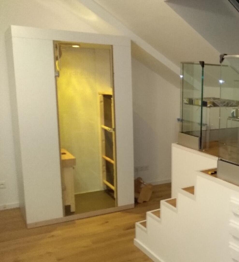 Eine kleine hölzerne Infrarotsauna mit einer Glastür ist in einem modernen Dachgeschossraum mit schrägen Decken, Holzböden und einer Treppe mit Glasgeländer in der Nähe installiert.