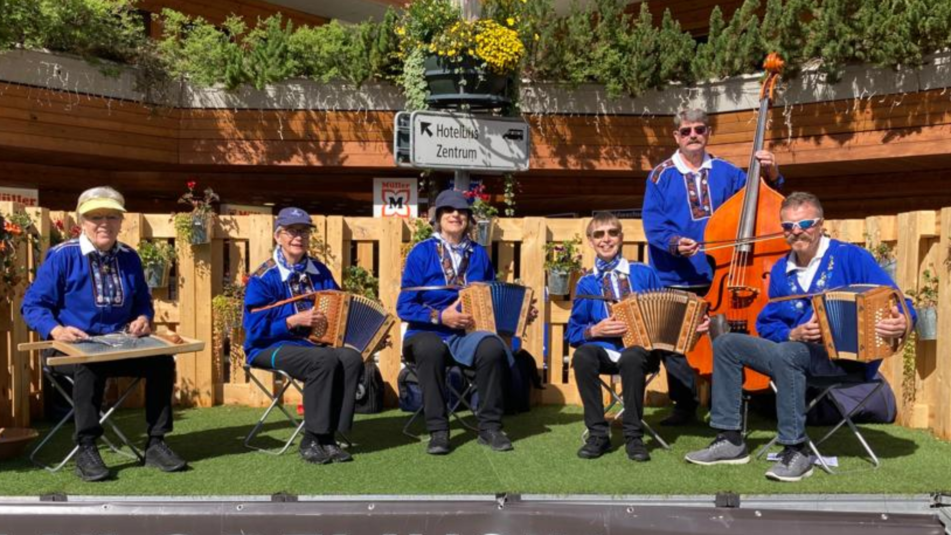 Sechs Musiker in passender blauer Kleidung spielen im Freien mit traditionellen Instrumenten, darunter Akkordeons, ein Kontrabass und ein Hackbrett, vor einer mit Blumen geschmückten Holzkulisse.