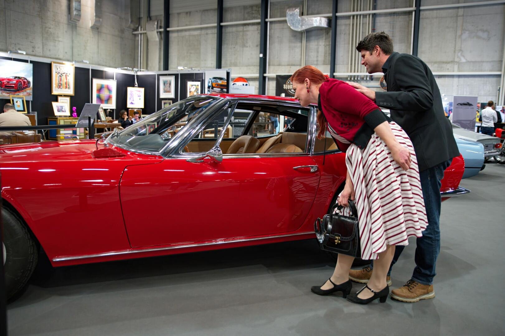 Eine Frau in einem rot-weißen Kleid und ein Mann in einem dunklen Blazer beugen sich vor, um einen Blick in einen glänzenden roten Oldtimer zu werfen, der auf einer Autoausstellung mit gerahmter Kunst an den Wänden im Hintergrund steht.