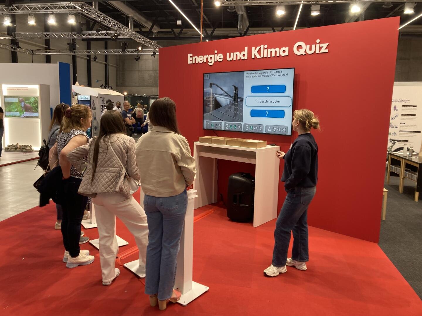 Fünf Personen stehen vor einer großen roten Wand mit der Aufschrift "Energie und Klima Quiz" und beschäftigen sich mit einem interaktiven Quiz, das auf einem Bildschirm bei einer Indoor-Veranstaltung oder Ausstellung angezeigt wird.