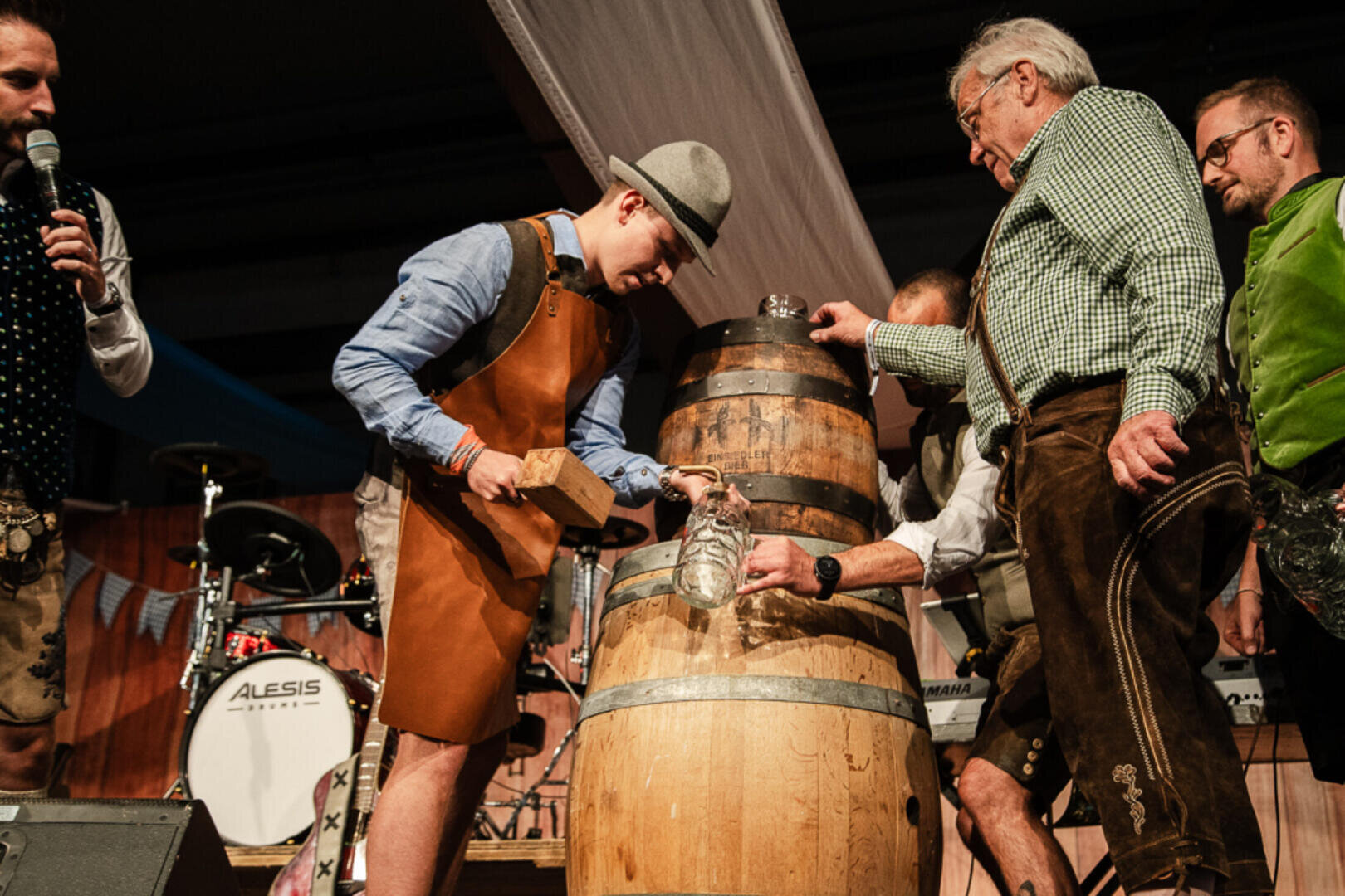 Eine Gruppe von Männern in traditioneller bayerischer Kleidung versammelt sich um ein hölzernes Bierfass auf der Bühne; ein Mann zapft das Fass an, während andere Krüge halten, und im Hintergrund sind Musikinstrumente zu sehen.