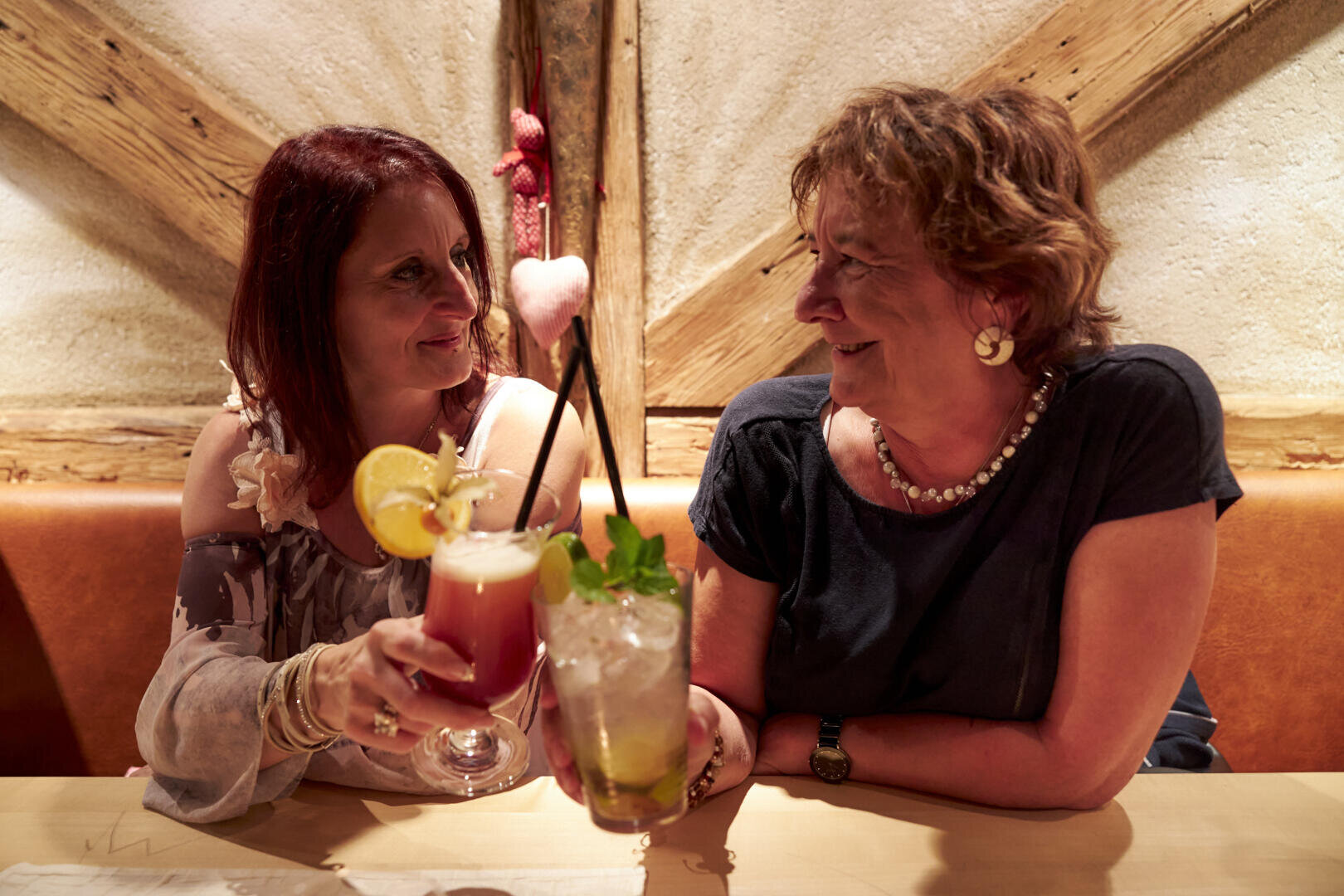 Zwei Frauen sitzen an einem Tisch in einem gemütlichen Restaurant, lächeln sich an und halten bunte Cocktails in der Hand, als würden sie einen Toast aussprechen. Die warme Beleuchtung und das rustikale Holzdekor schaffen eine fröhliche, freundliche Atmosphäre.