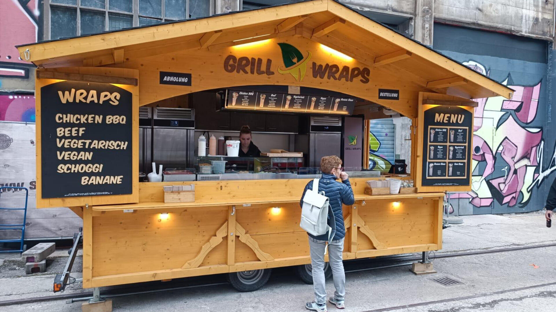 Eine Person steht vor einem gelben Holzgrill, an dem ein Imbissstand mit Speisekarten aufgebaut ist, an dem BBQ-Hähnchen, Rindfleisch, vegetarische und vegane Gerichte sowie Schokoladen- und Bananen-Wraps angeboten werden. Der Stand steht vor einer mit Graffiti besprühten Wand.