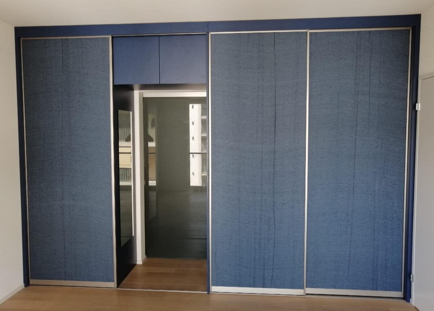 Ein moderner Kleiderschrank mit blauen Schiebetüren und Metallverzierungen ist teilweise geöffnet und gibt den Blick auf einen großen Spiegel im Inneren frei. Der Boden ist aus Holz und die Wände sind schlicht weiß.
