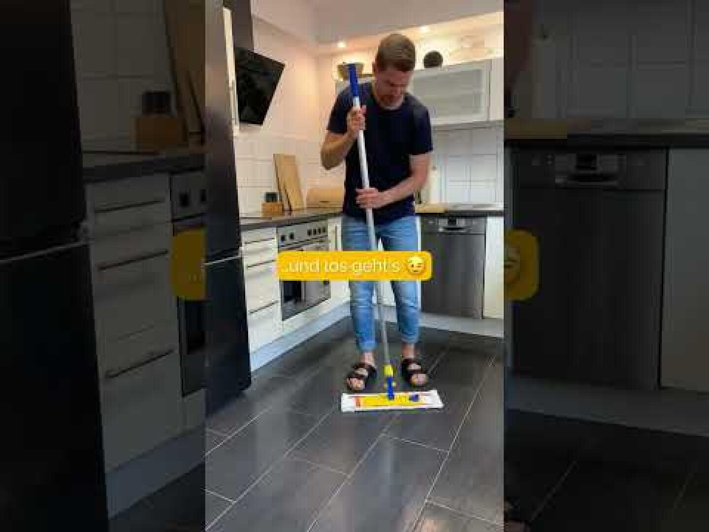 Eine Person, die ein marineblaues Hemd, Jeans und Sandalen trägt, wischt den Boden in einer modernen Küche. Eine gelbe Beschriftung mit einem Smiley auf Deutsch lautet "und los gehts".