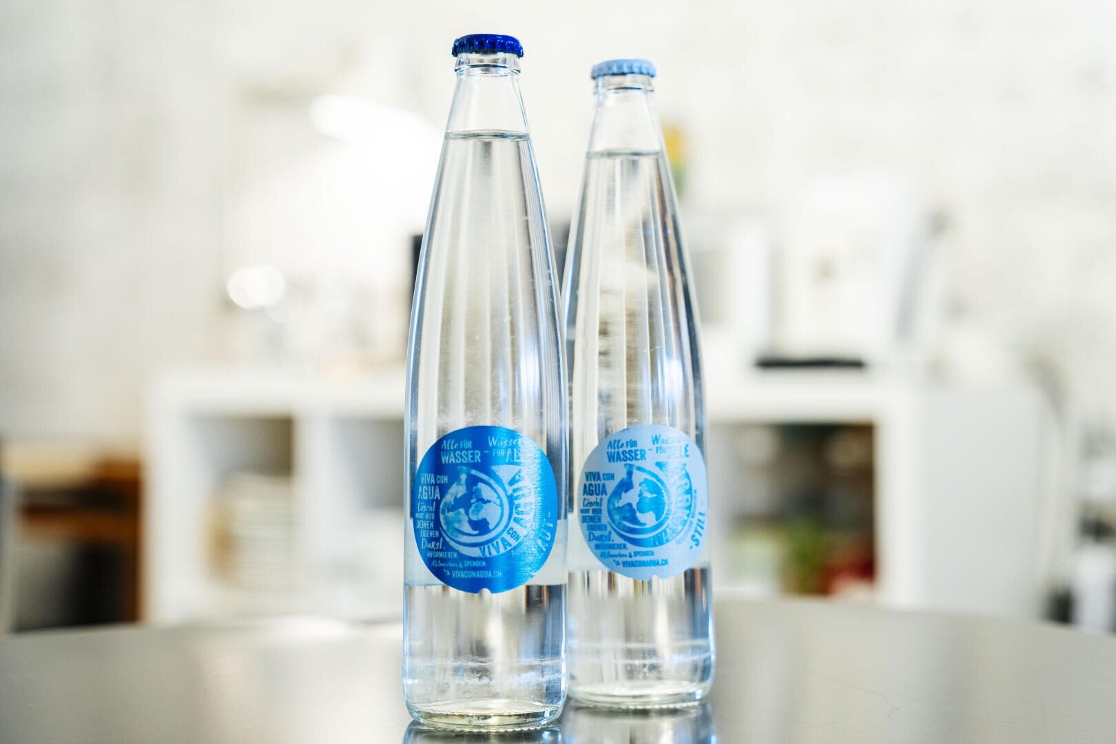 Zwei klare Glasflaschen mit blauen Etiketten stehen nebeneinander auf einer spiegelnden Oberfläche, vor einem verschwommenen Innenhintergrund mit weißen Regalen und verschiedenen Gegenständen.