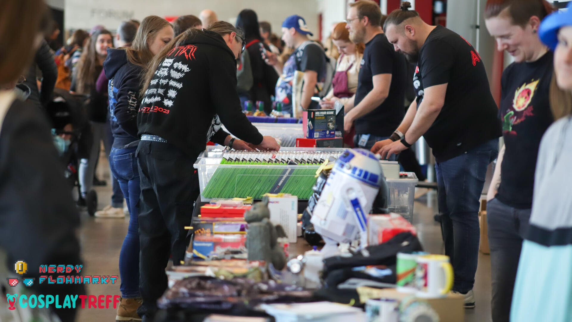 Menschen stöbern auf einer Messe oder einem Markt durch Tische mit verschiedenen Sammlerstücken, Büchern und Spielzeug. Einige Besucher tragen Cosplay- oder Themen-Shirts, und auf einem der Tische ist ein R2-D2-Modell zu sehen.