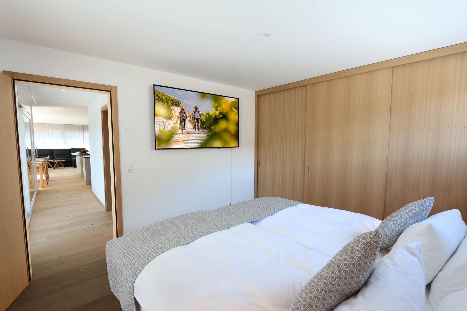 Ein modernes Schlafzimmer mit einem ordentlich gemachten Bett, gemusterten Kissen, großen Holzschränken und einem an der Wand montierten Fernseher, der die Menschen zeigt, die die Treppe hinaufgehen. Eine offene Tür führt in einen hellen Wohn- und Essbereich mit Holzböden.
