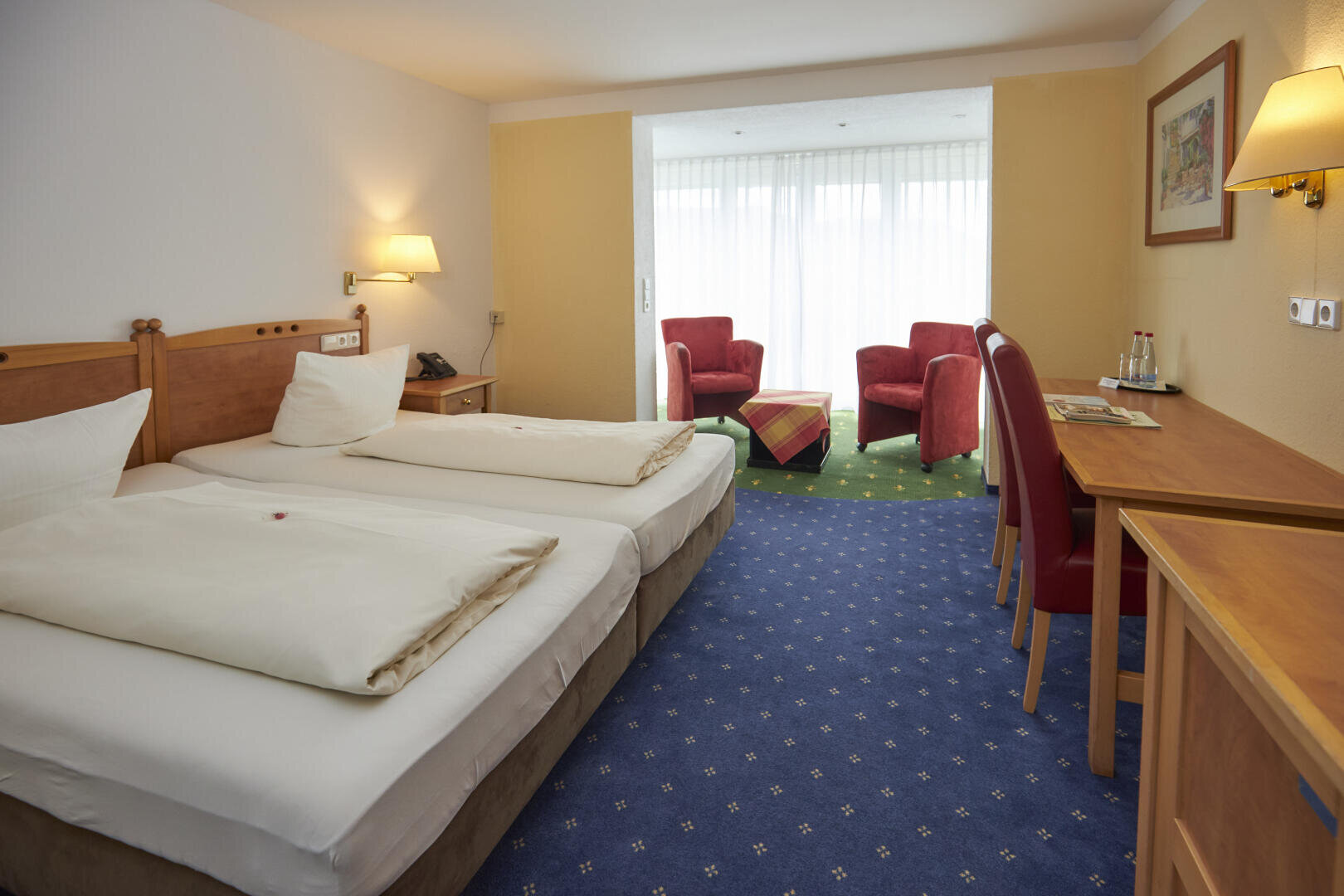 Hotelzimmer mit zwei zusammengeschobenen Einzelbetten, einem Holztisch mit Stühlen, einer Wandlampe, einem Gemälde und einer Sitzecke mit zwei roten Sesseln und einem Tisch an großen Fenstern mit durchsichtigen Vorhängen.