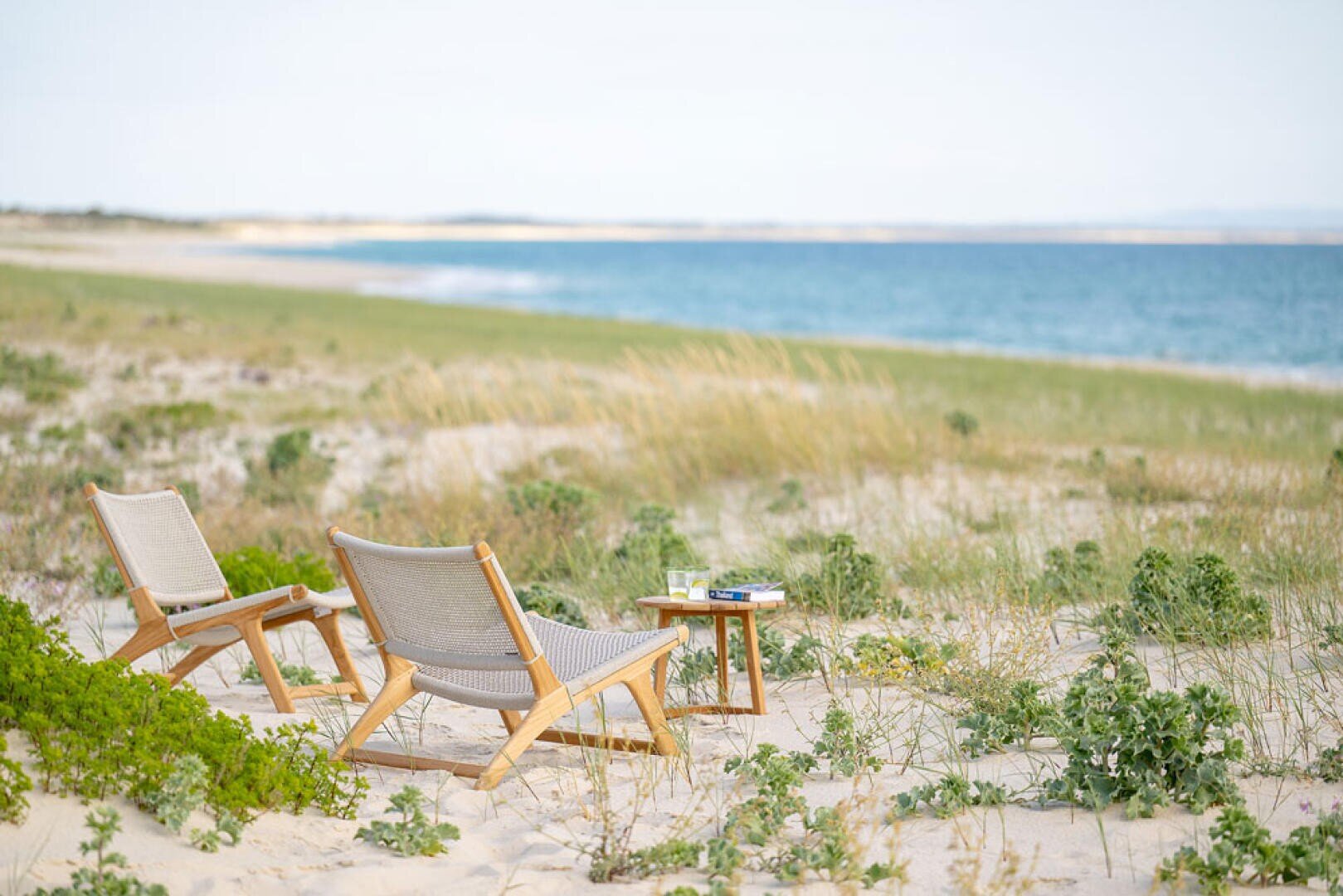 Zwei hölzerne Liegestühle und ein kleiner Beistelltisch mit Büchern stehen auf einem Sandstrand mit grünem Gras und Pflanzen, mit Blick auf das ruhige blaue Meer unter einem klaren Himmel.
