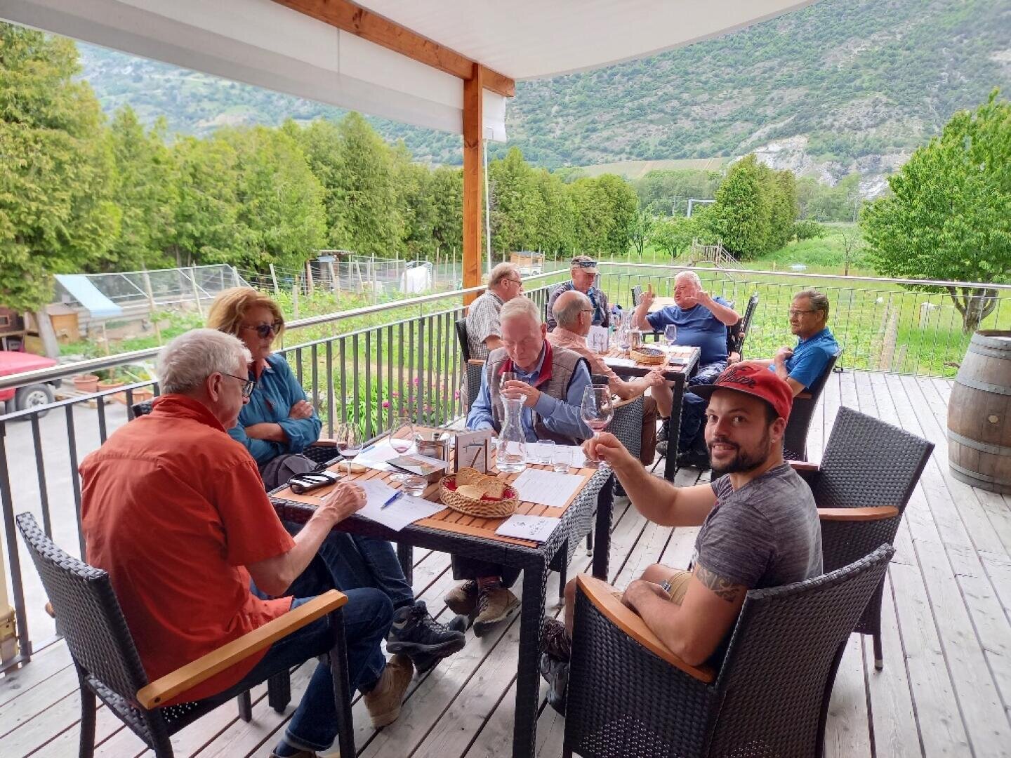 Eine Gruppe von Menschen sitzt um einen Tisch auf einer Holzterrasse im Freien und genießt Essen und Getränke. Grüne Hügel und Bäume umgeben die Terrasse und schaffen eine friedliche, malerische Kulisse. Eine Person im Vordergrund zeigt den Daumen nach oben.