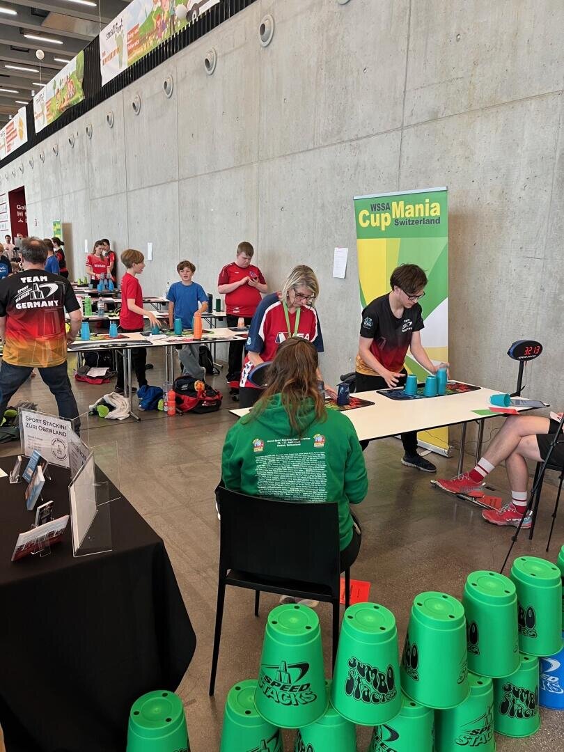 Mehrere Personen nehmen an einem Becherstapelwettbewerb in einer großen Halle teil. Im Vordergrund sind grüne Plastikbecher gestapelt, und es sind Banner für "WSSA Cup Mania Switzerland" zu sehen.
