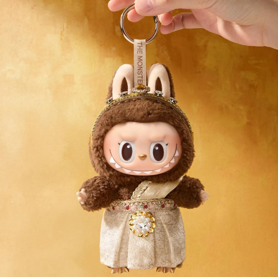 Eine Hand hält einen Schlüsselanhänger, auf dem eine niedliche Puppe mit großen Augen, Hasenohren und einem pelzigen braunen Anzug abgebildet ist. Die Puppe trägt eine verschnörkelte goldene Schärpe und eine Krone vor einem warmen goldenen Hintergrund.