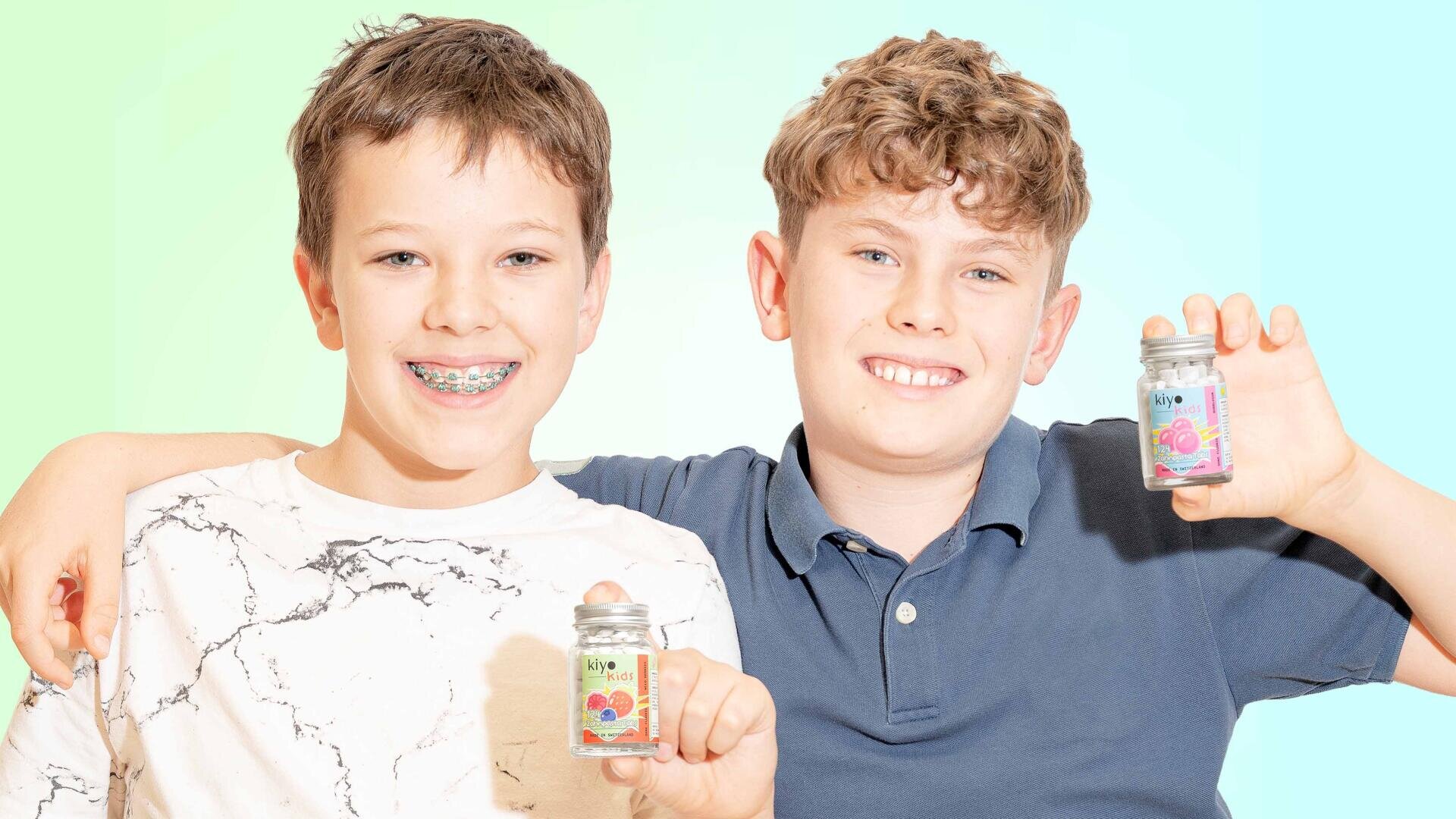 Zwei lächelnde Jungen stehen dicht beieinander und halten jeweils ein kleines Glas mit bunten Etiketten und Süßigkeiten darin hoch. Der Junge auf der linken Seite hat eine Zahnspange und trägt ein weißes Hemd; der Junge auf der rechten Seite hat lockiges Haar und trägt ein blaues Poloshirt.
