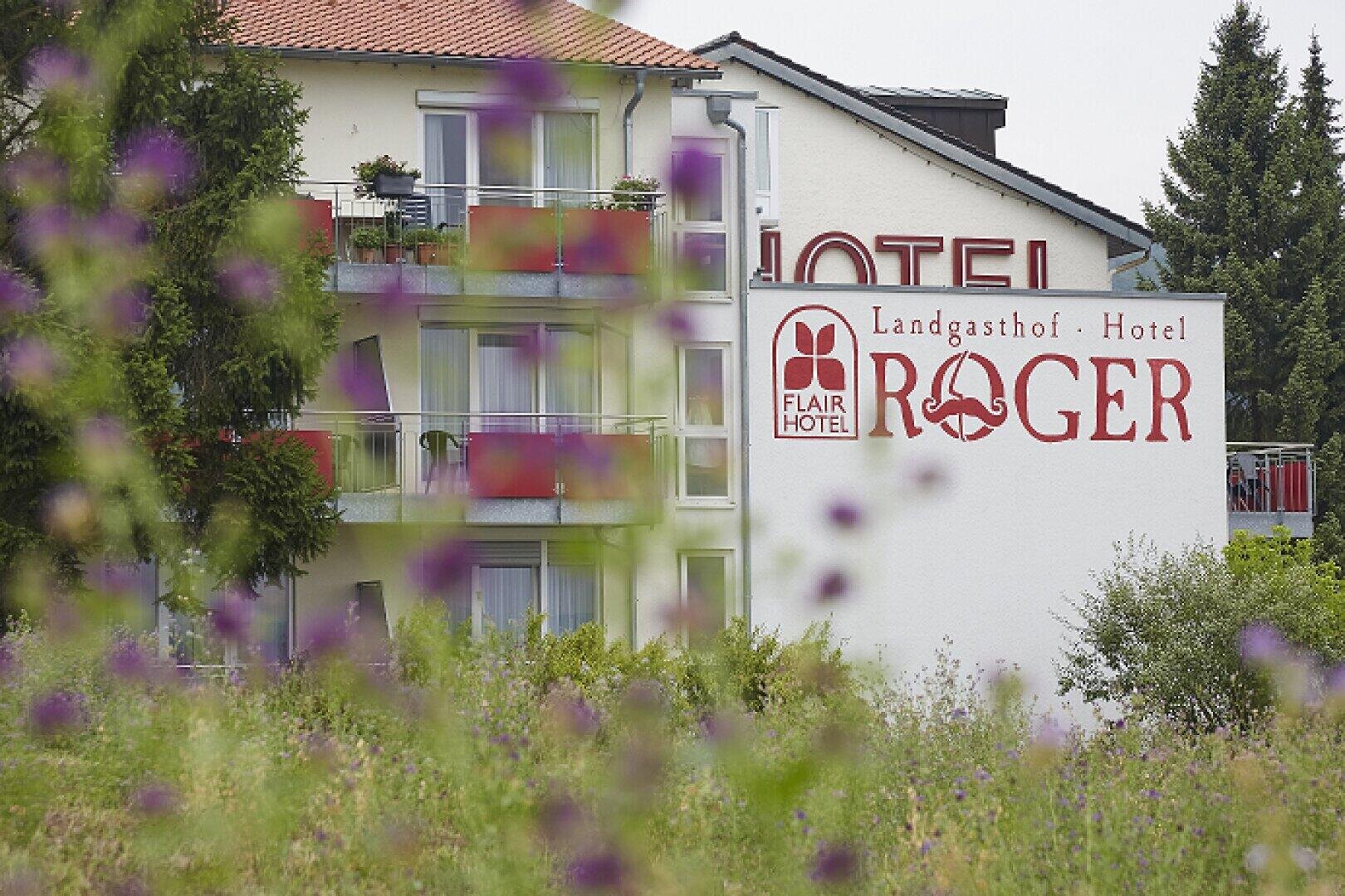 Ein Hotelgebäude mit Balkonen wird teilweise von Wildblumen im Vordergrund verdeckt. Auf einem großen Schild an der Wand steht Landgasthof Hotel Roger mit dem Logo des Flair Hotels. Im Hintergrund sind Bäume zu sehen.