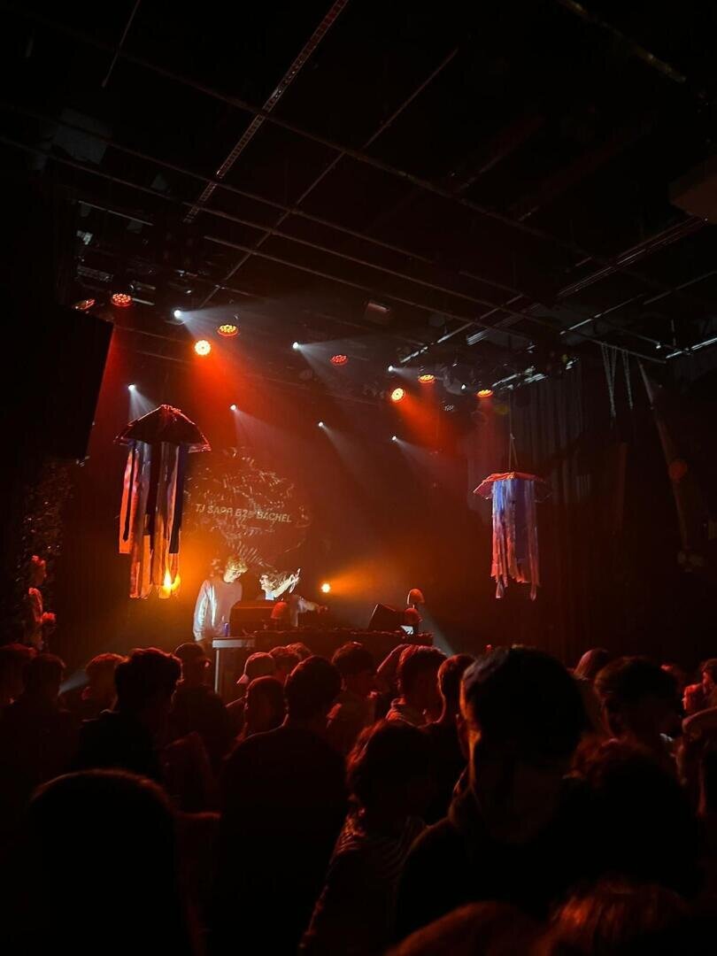 Eine Menschenmenge steht vor einer Bühne, auf der zwei Musiker unter roten und orangefarbenen Lichtern spielen. Von der Decke hängen quallenförmige Dekorationen, und an der Rückwand ist eine kreisförmige Projektion zu sehen.